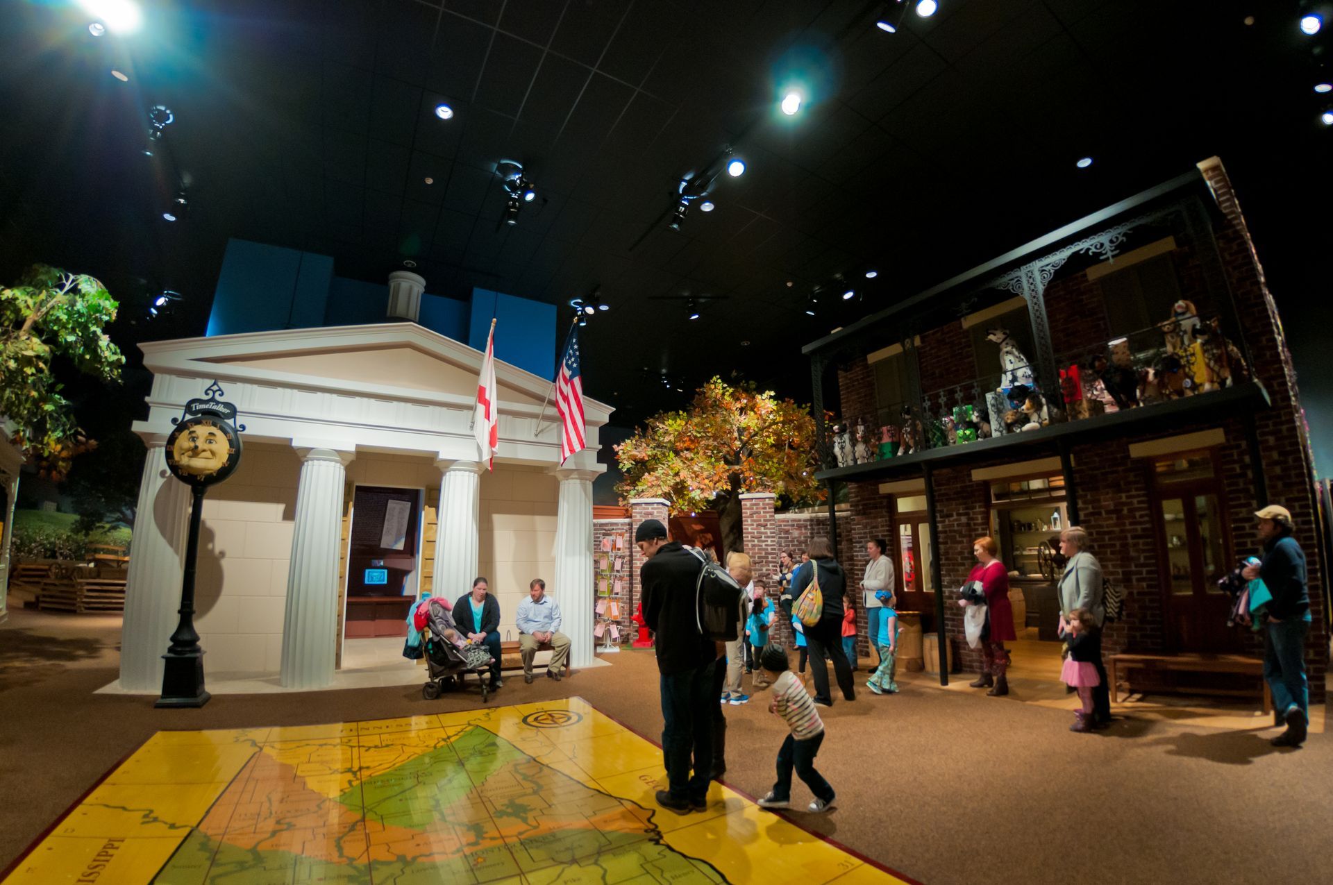 Museum exhibit: Replica town square with people viewing displays. Buildings are cream and brick, with a large map on the floor.