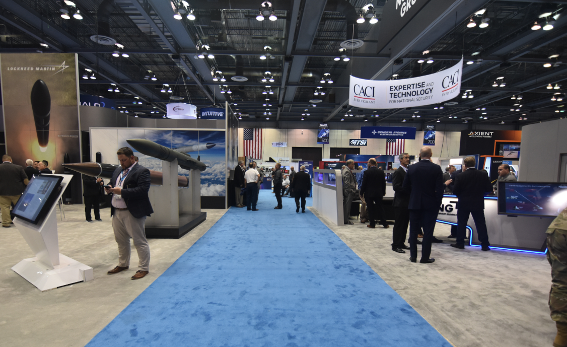 People at a trade show with booths and displays. A blue carpeted aisle leads through the center.