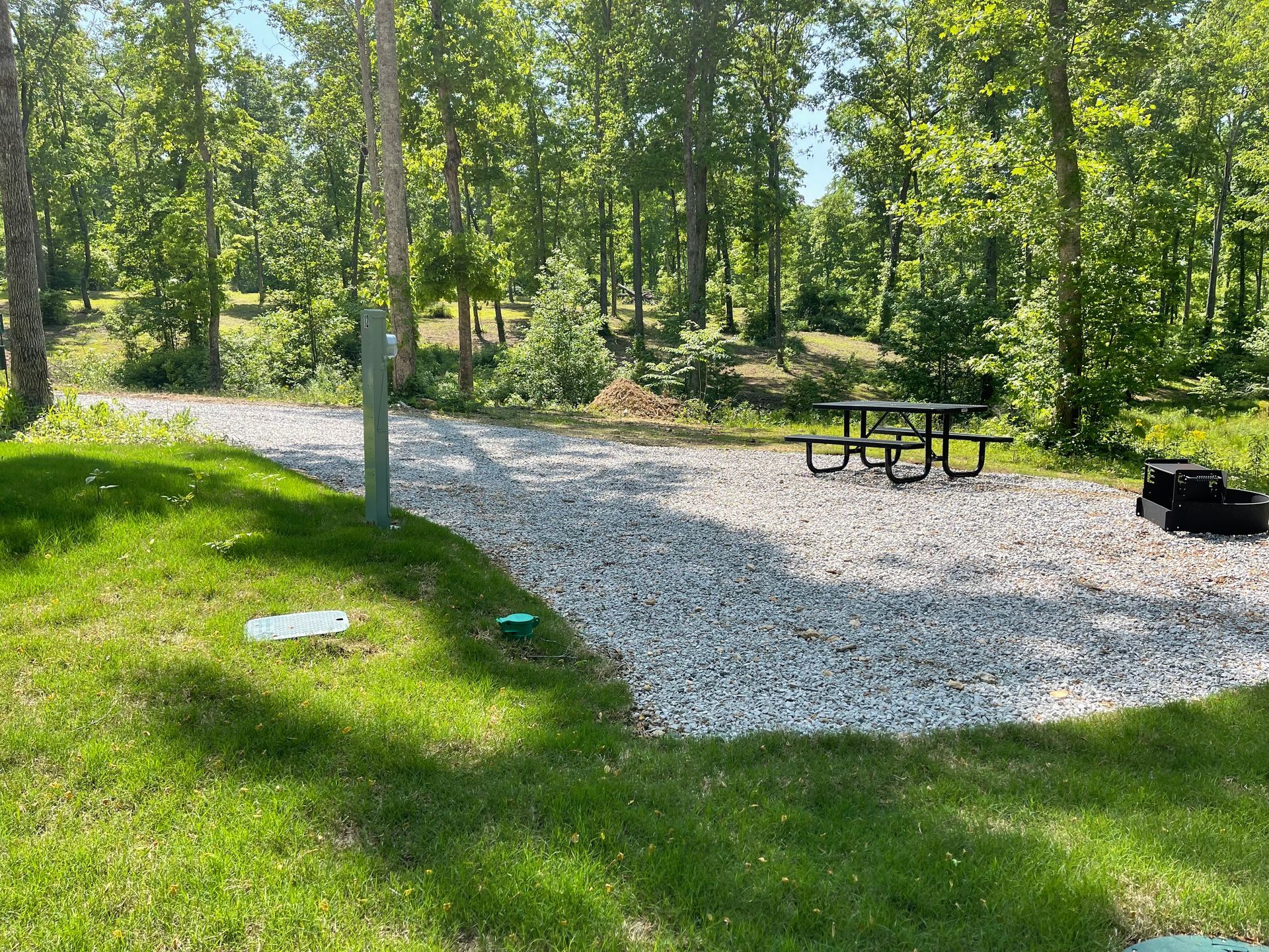 A gravel camping site with a picnic table, fire pit, and electrical hookup in a wooded area.