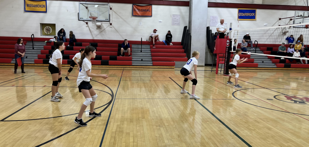 Indoor volleyball game in a gym, players in white and black uniforms poised near the net