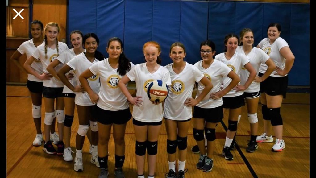 Volleyball team posing in white jerseys and black shorts inside a gym, hands on hips, smiling.