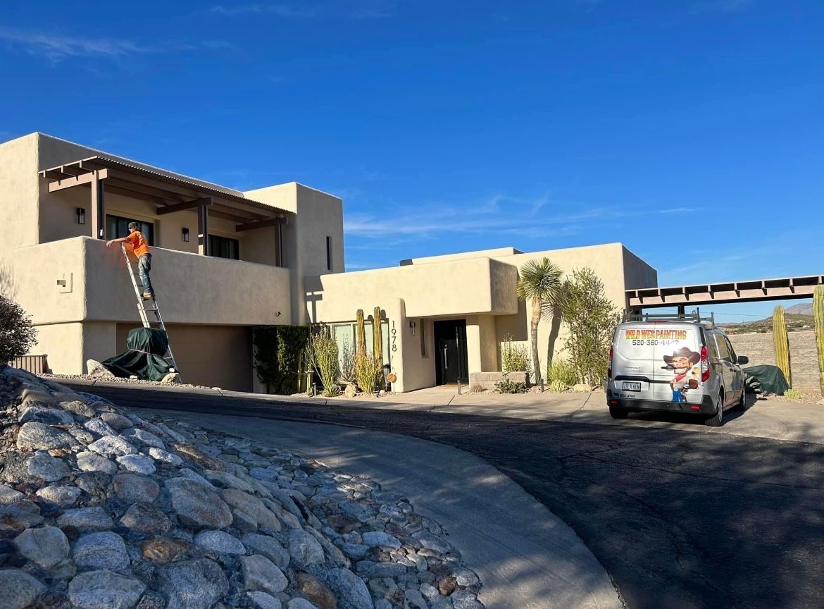 A person on a ladder works on a tan stucco home. A van with a company logo is parked nearby, under a bridge.