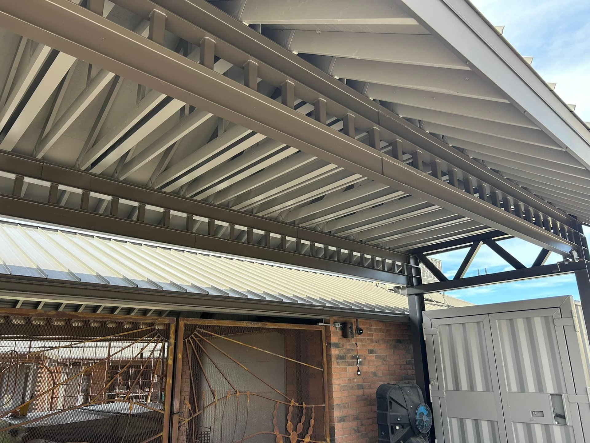 A carport with a brown metal frame and roof over a light-colored garage door and brick wall.