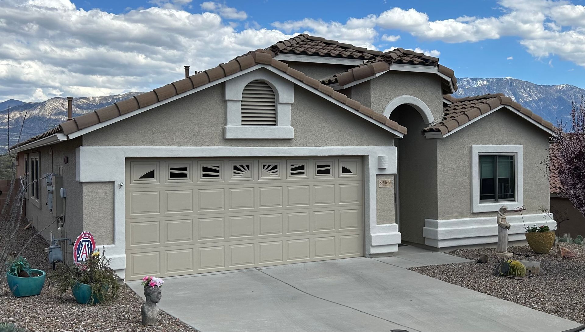 Tan stucco house with a beige garage door, set against a backdrop of mountains and a partly cloudy sky.