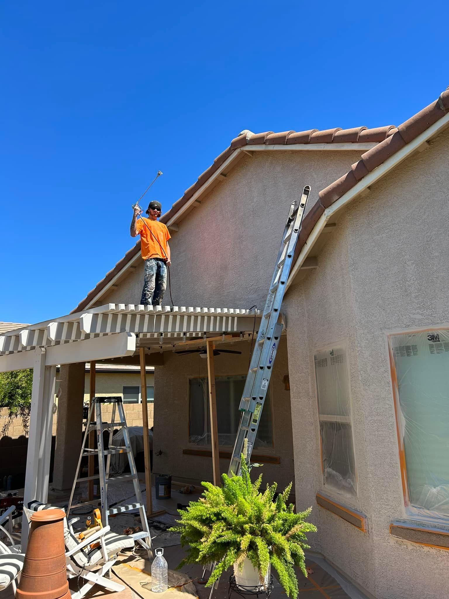 A person in an orange shirt on a ladder working on the roof of a house with a pergola and blue sky in the background.