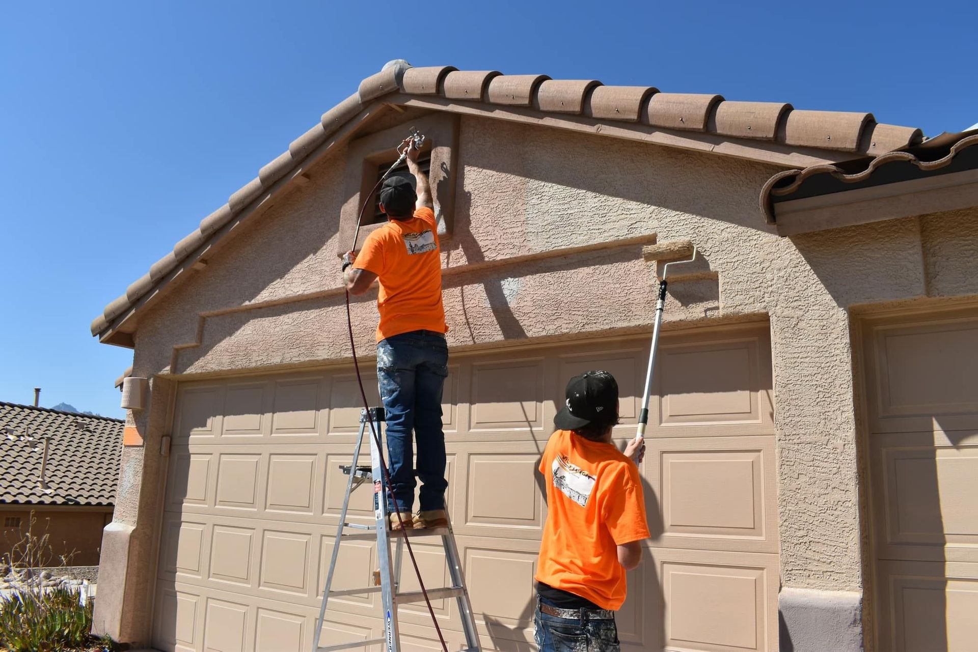 Two workers in orange shirts painting a tan garage under a blue sky. One is on a ladder, the other uses a roller.