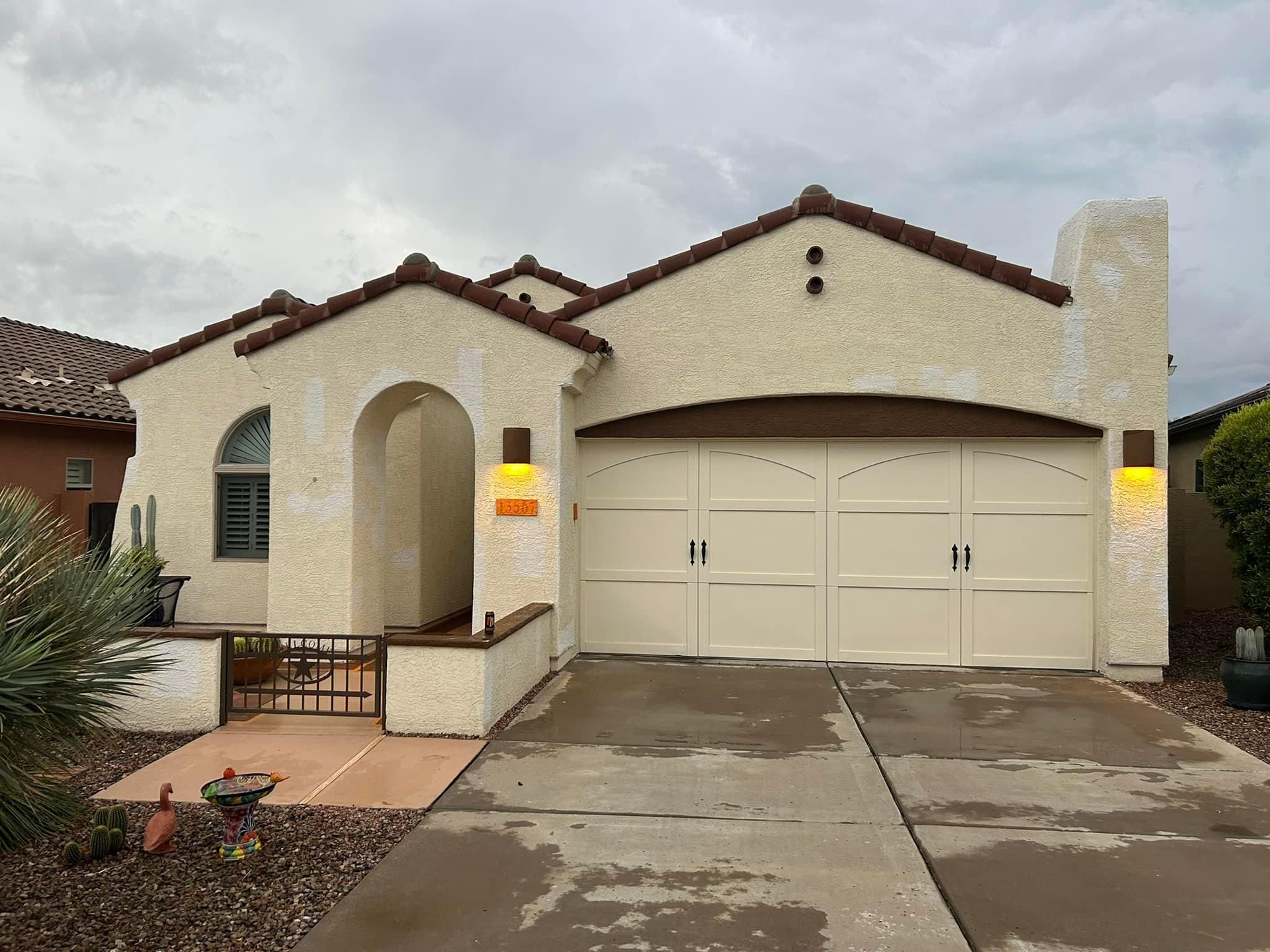 Beige stucco house with a curved archway, a two-car garage, and a terracotta tile roof, set against a cloudy sky.