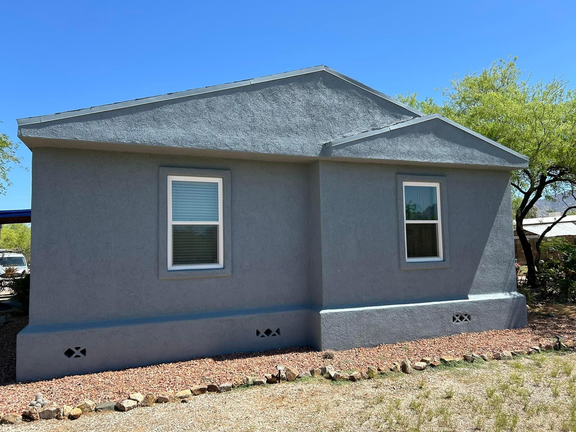 Gray stucco house with two windows, set against a blue sky. The house sits on a brown rock base with a small yard.