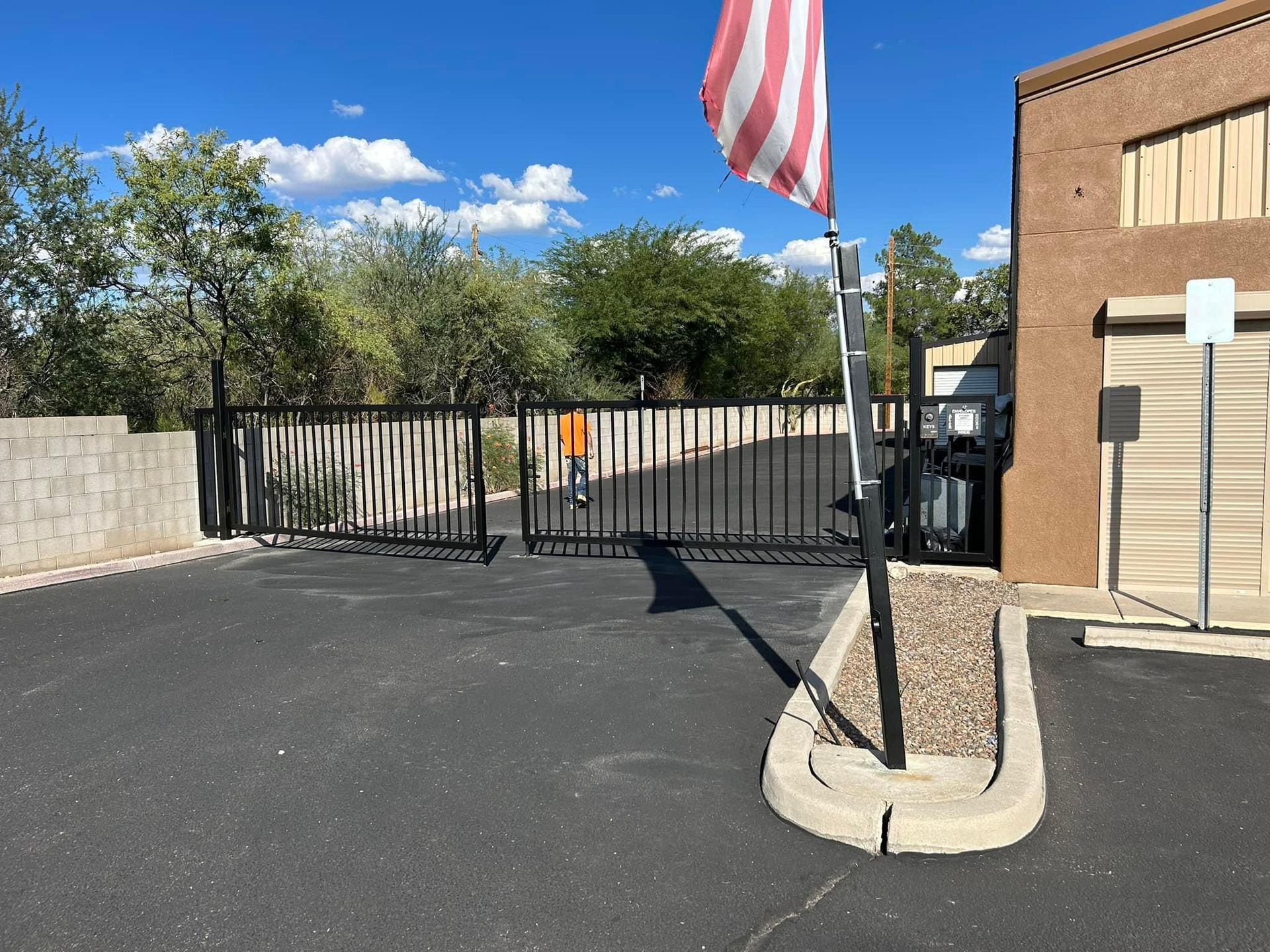 Black gated entrance with American flag waving. The gate is set against a building and a clear blue sky.