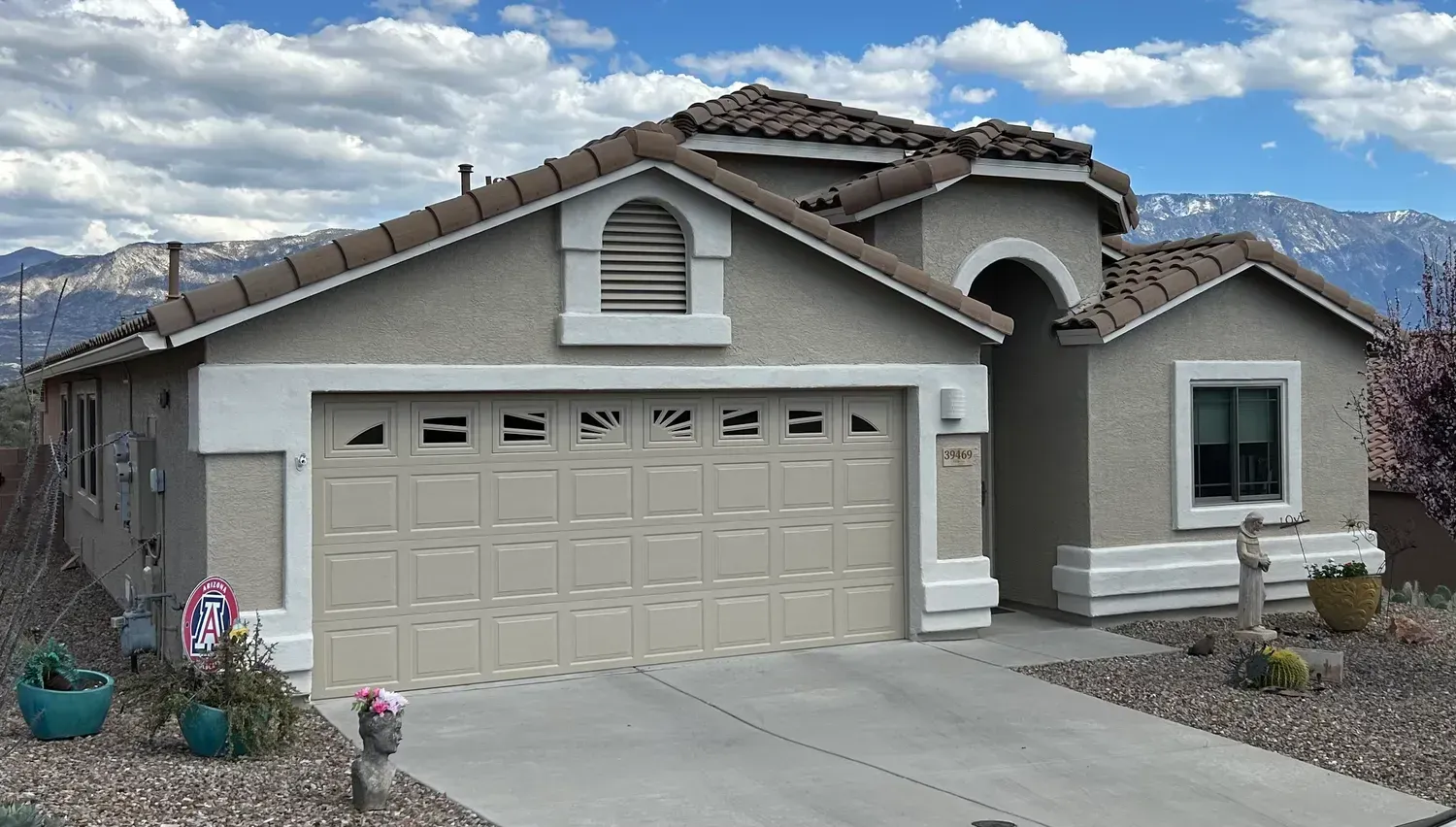Tan stucco house with a brown tile roof, beige garage door, and mountain backdrop under a cloudy sky.
