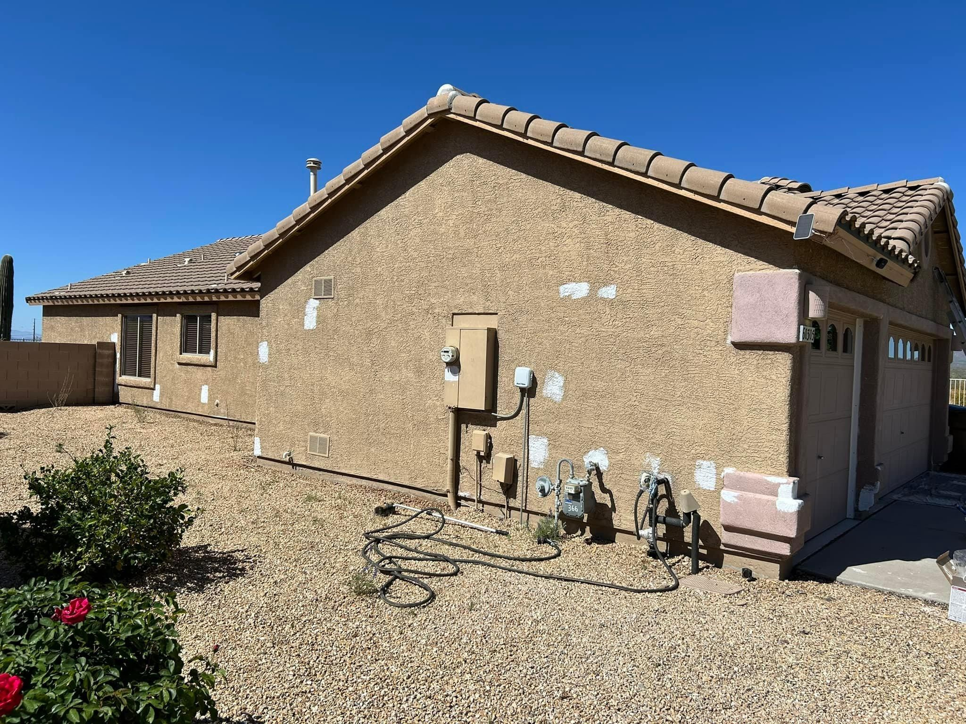 Tan stucco house with patches, electrical box, gas meters, and a gravel yard under a blue sky.