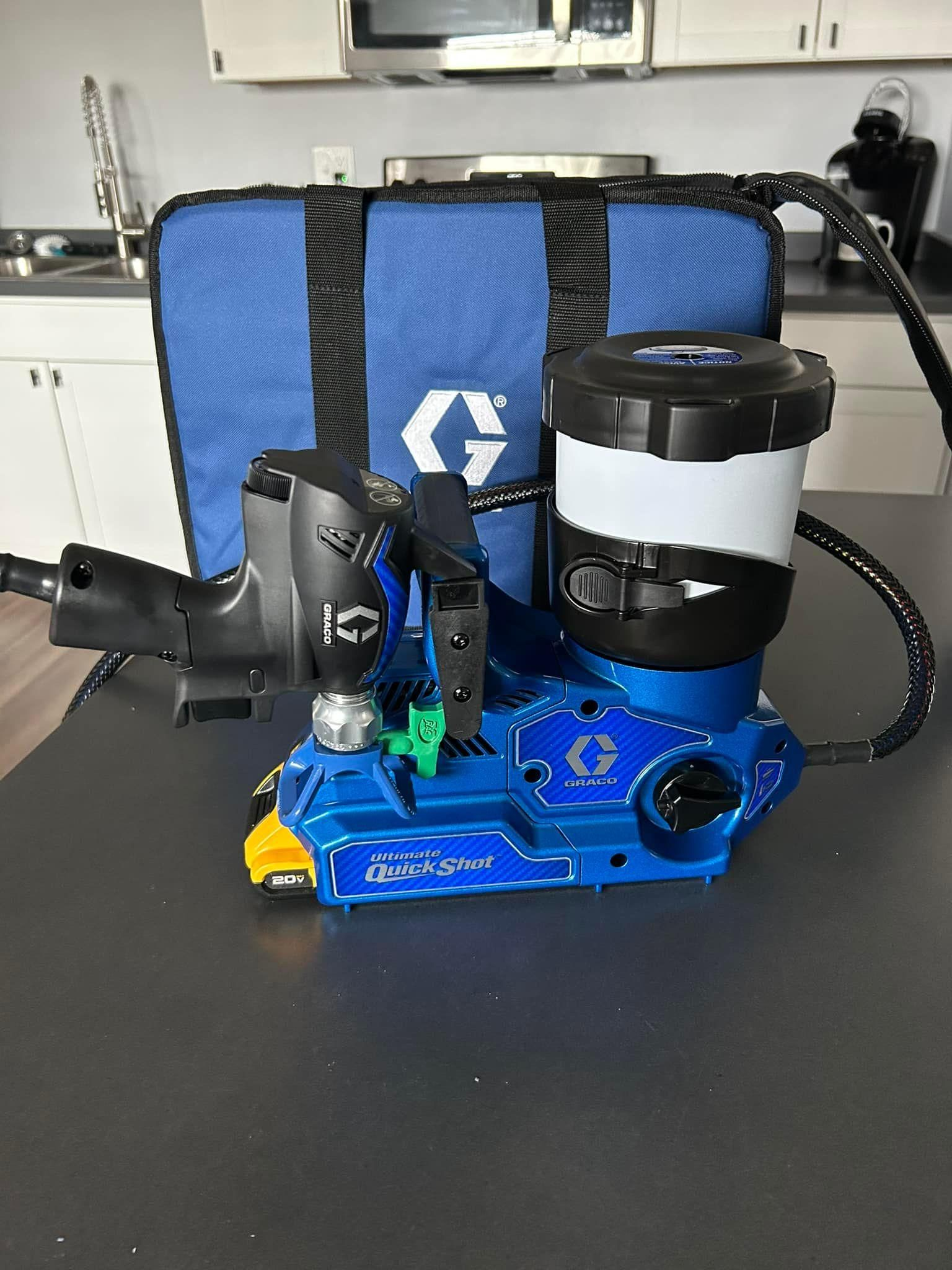 Blue Graco paint sprayer with a blue carrying bag on a dark countertop in a kitchen.