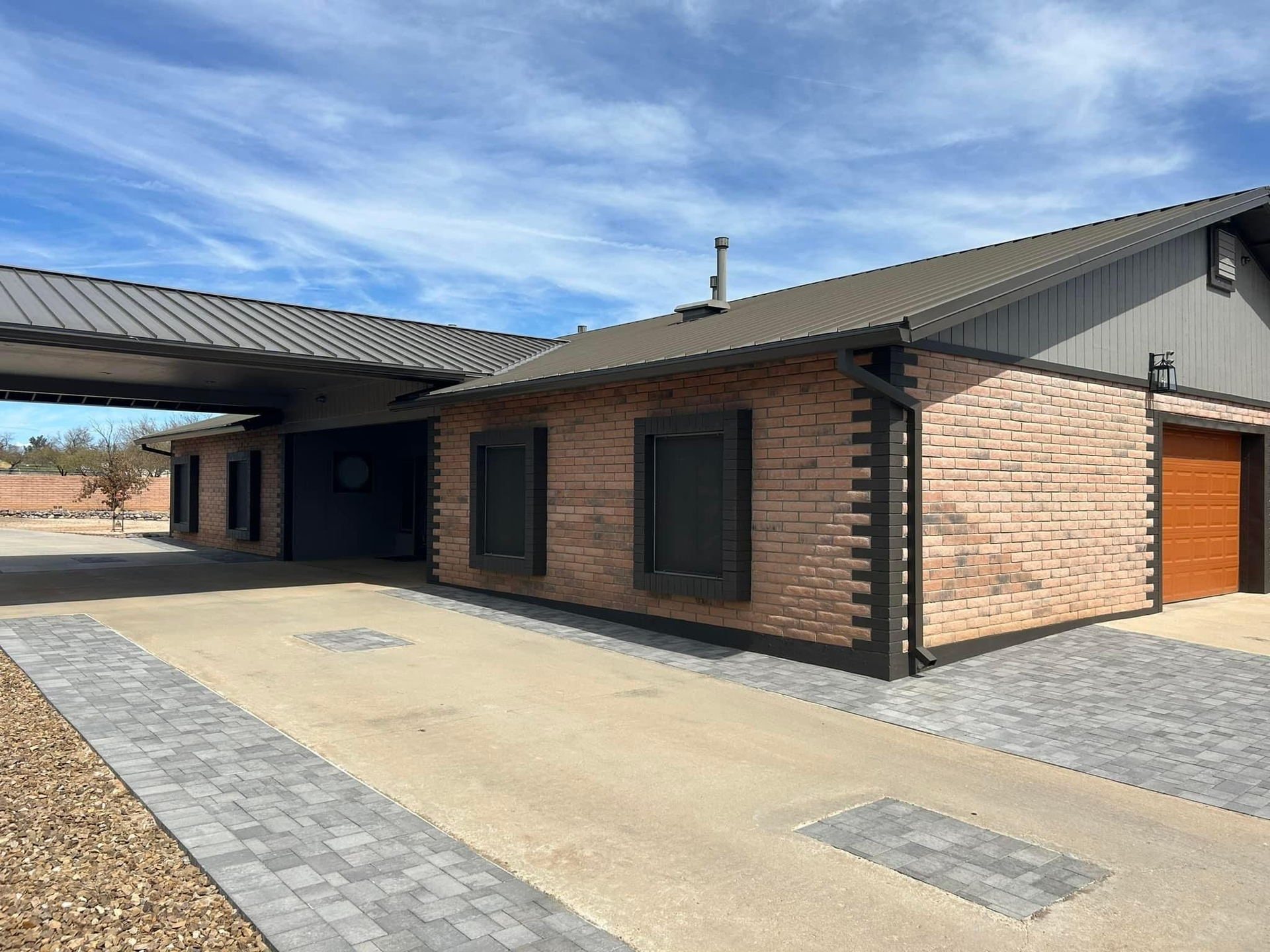 Exterior of a brick building with dark trim, a covered carport, and a paved driveway, under a blue sky.