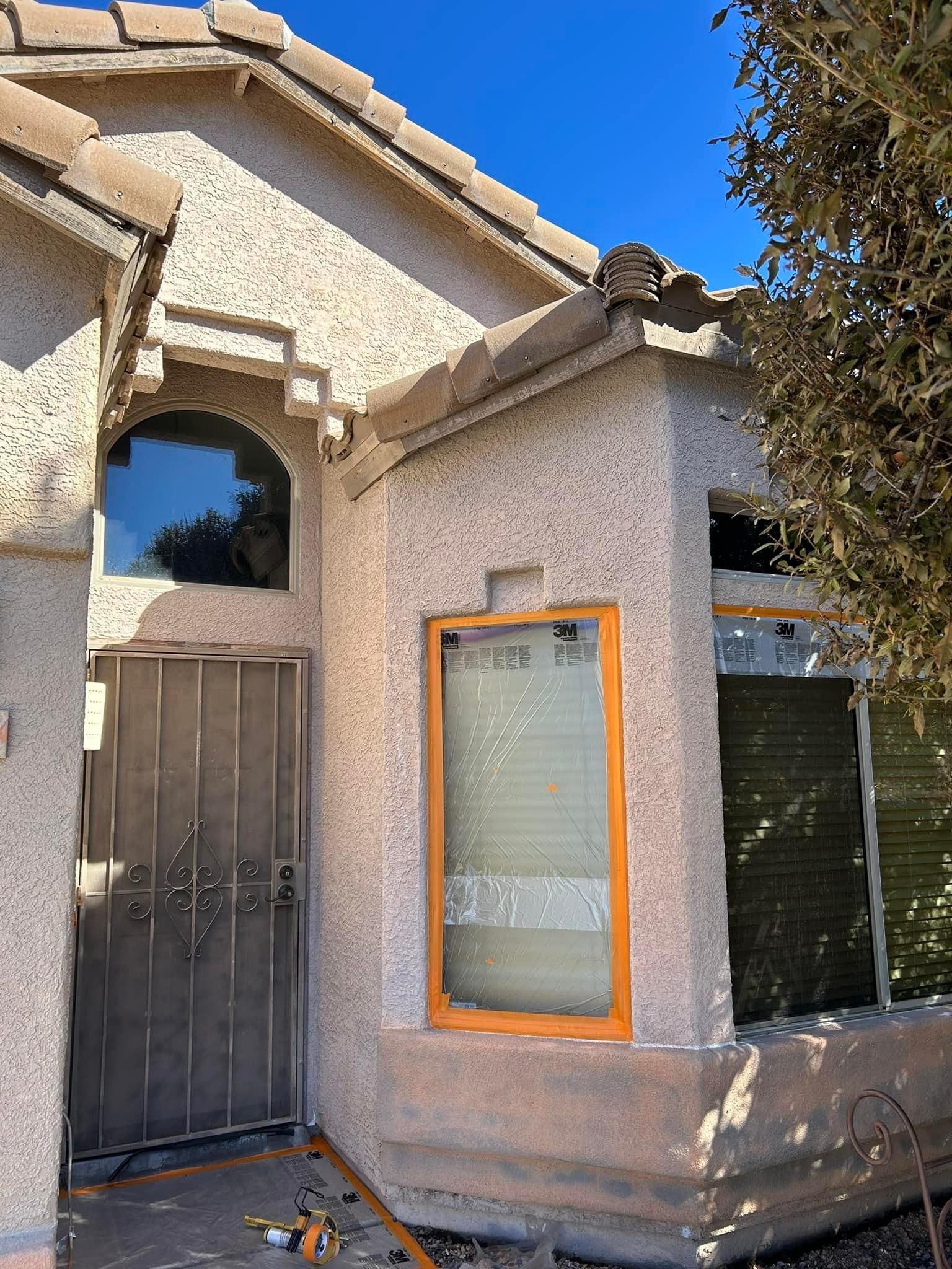 Exterior view of a stucco house with an arched window, security door, and a window with tan frame and blinds.
