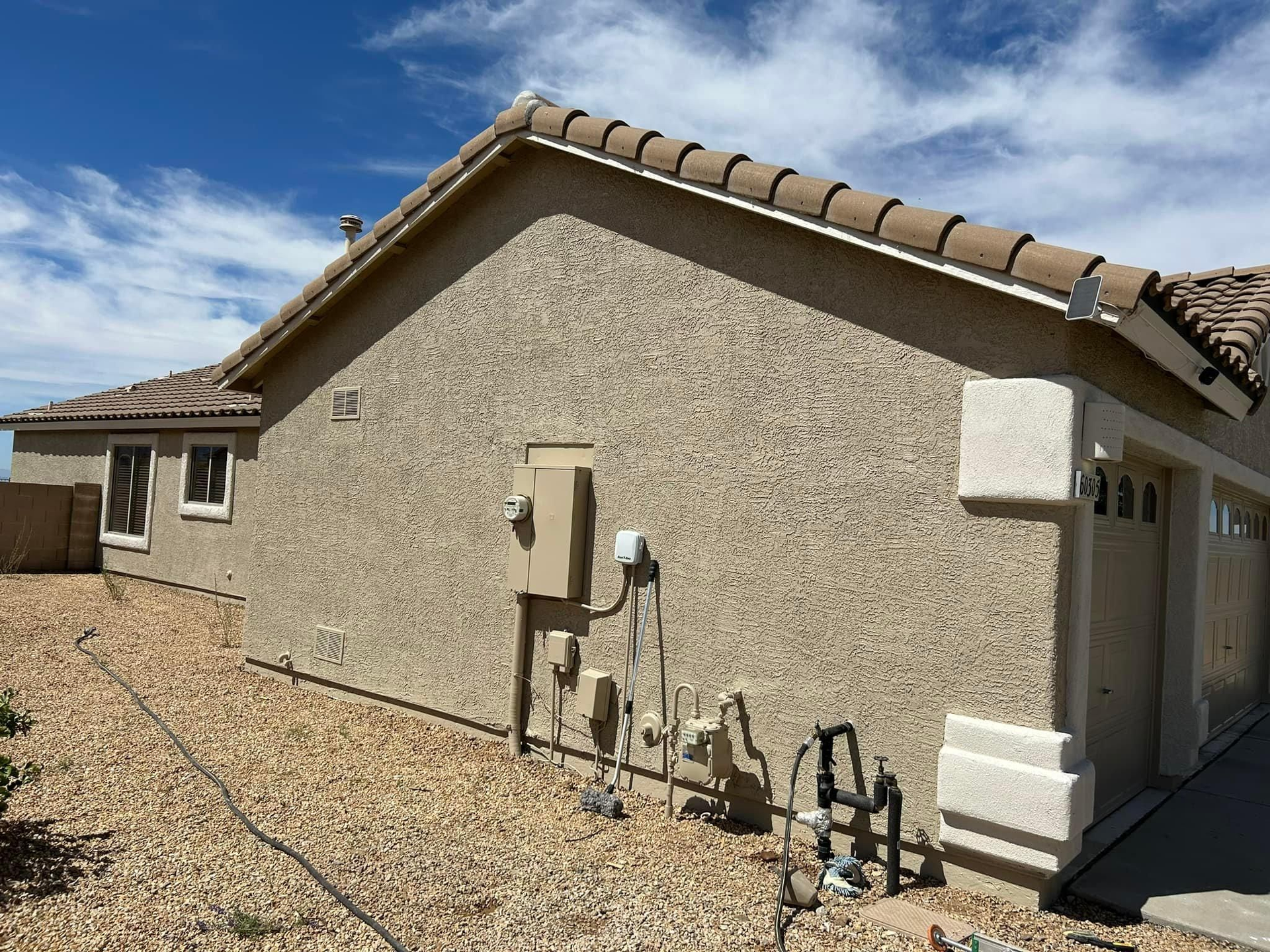 Tan stucco house with brown tile roof, electrical box, and gravel yard under a blue sky.