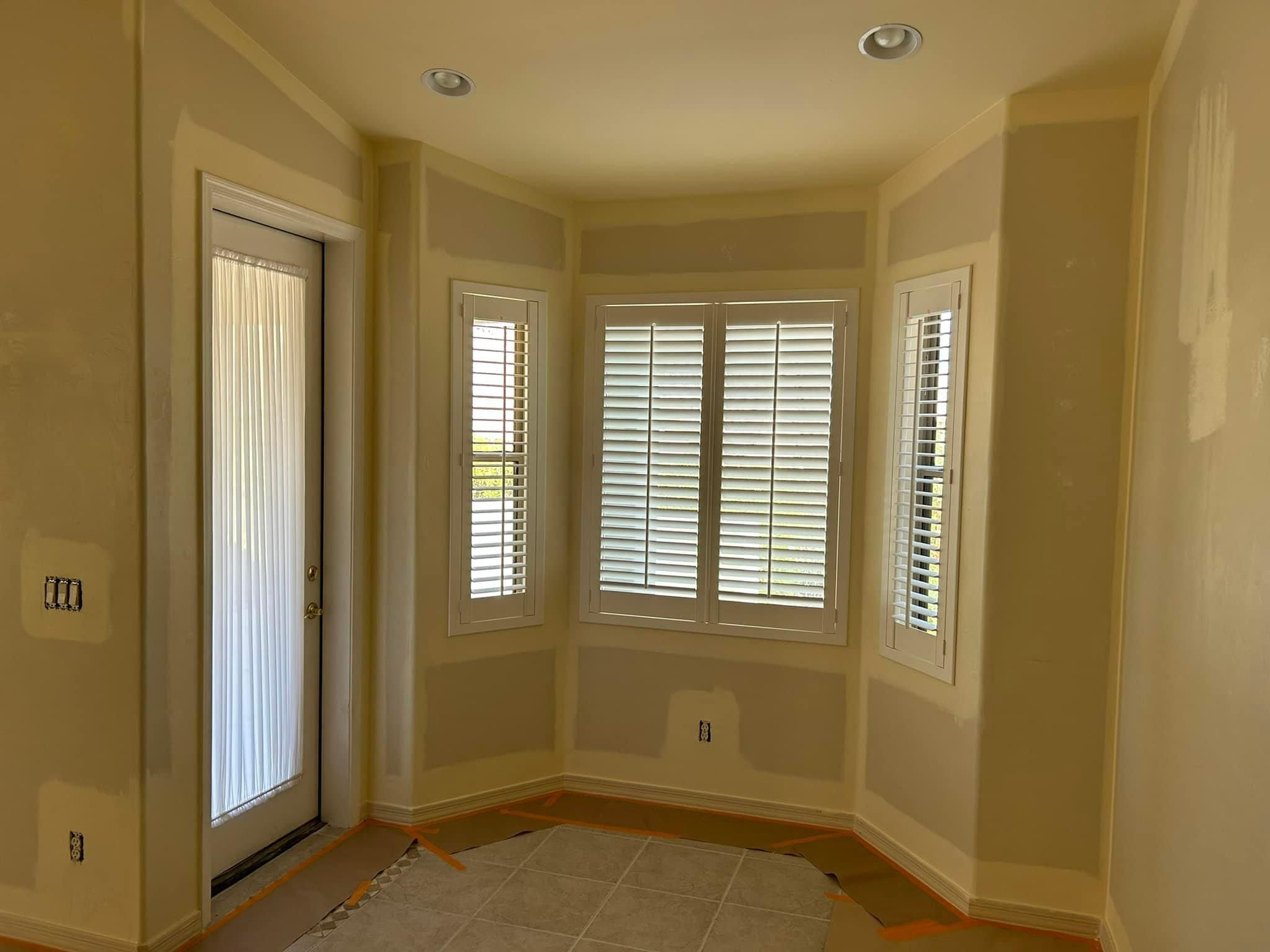 Interior view of a room with a door and bay window with shutters. Walls are light yellow with white trim.