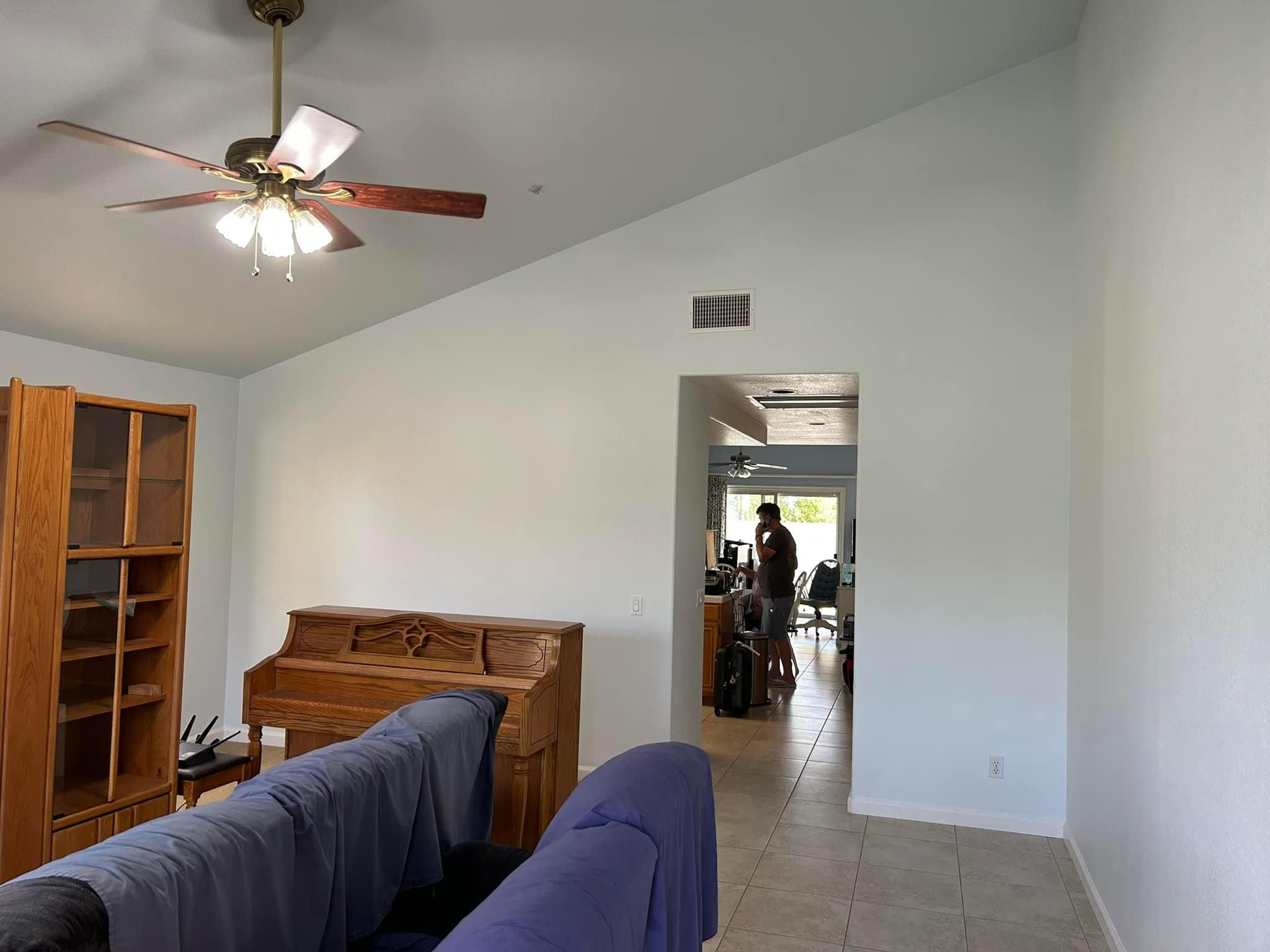 Living room with light blue walls, a piano, and a person vacuuming in the doorway. Brown wooden ceiling fan.