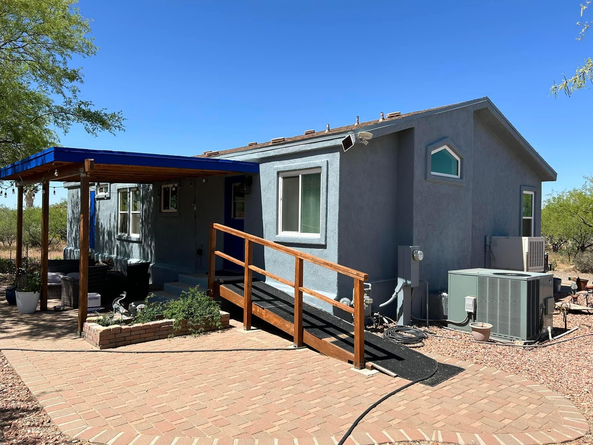 Small blue house with a wooden ramp leading to the entrance, under a blue awning. A brick patio is in front.