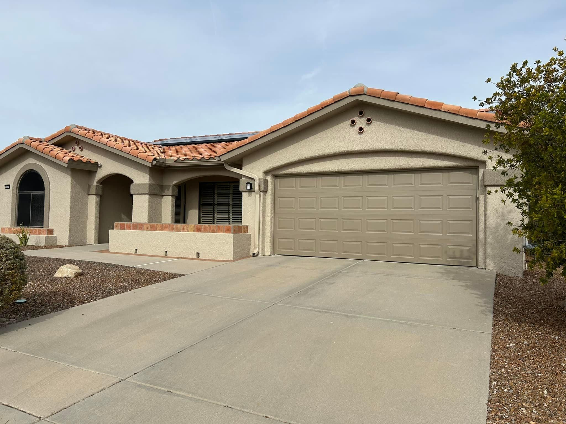 A light brown stucco house with a terracotta tile roof, garage, and driveway. Landscaping includes rocks and a small tree.