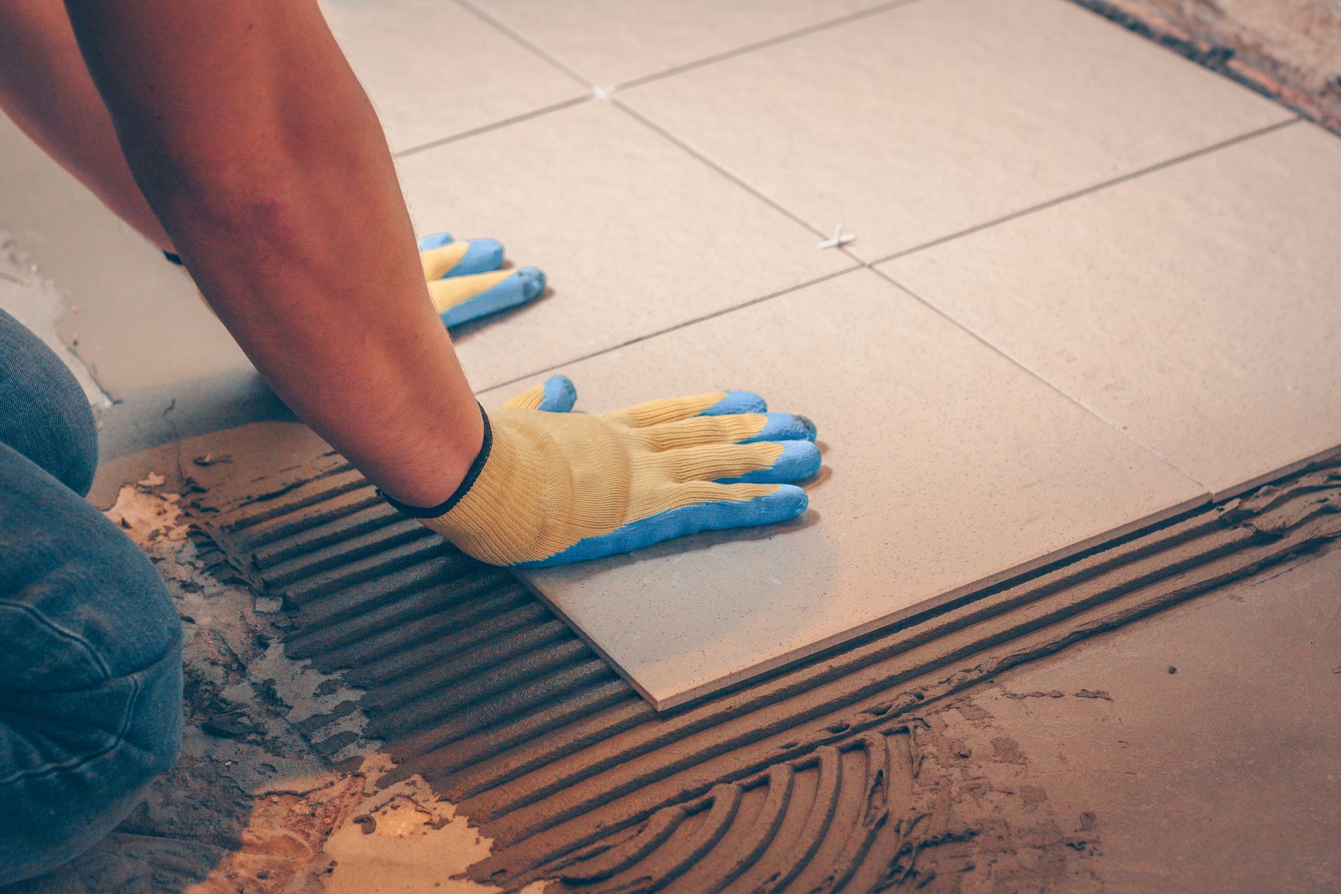 A person wearing blue and yellow work gloves presses a large square tile into thin-set mortar on a floor.