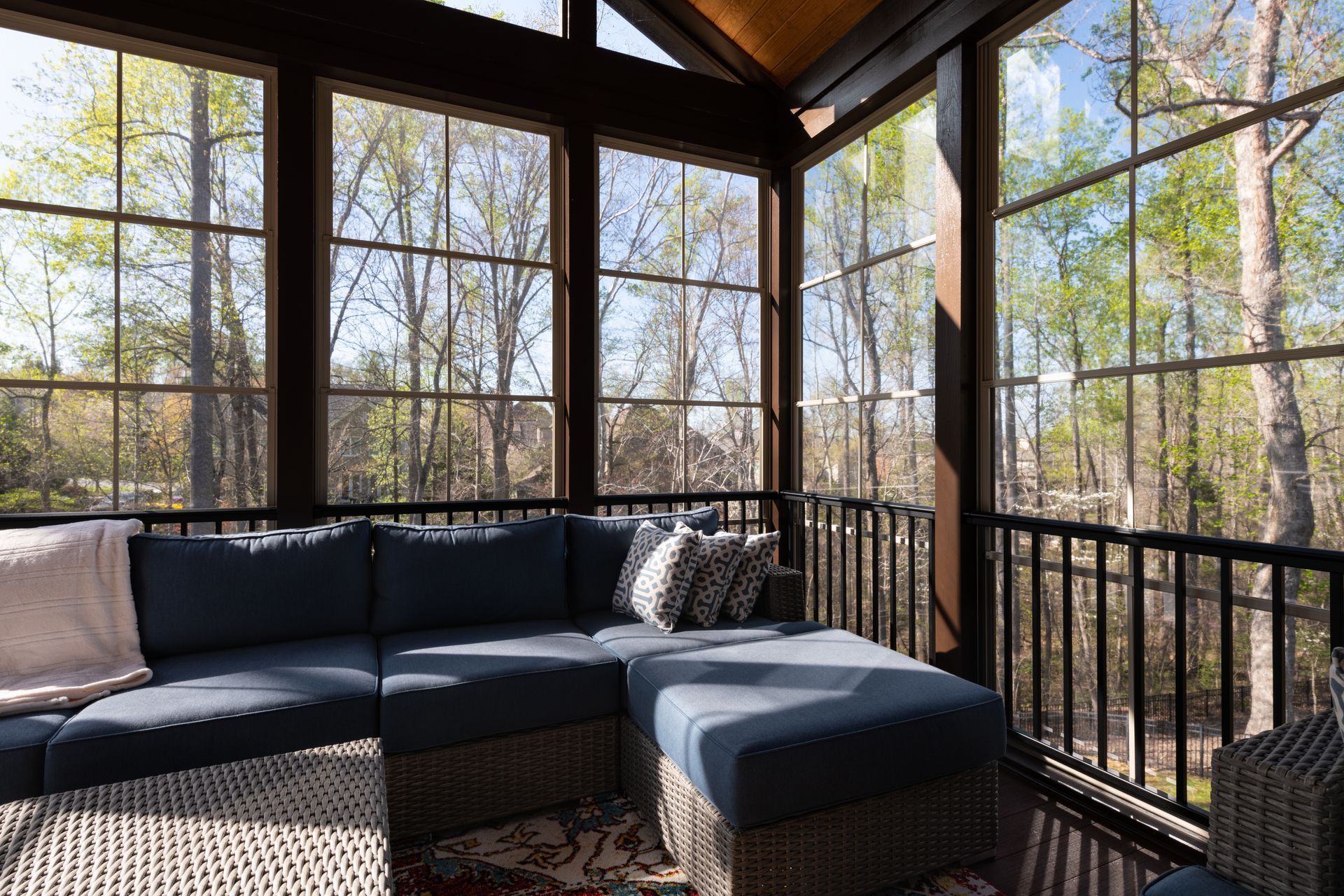 A blue sectional sofa sits on a rug in a sunroom with floor-to-ceiling windows overlooking a wooded area.