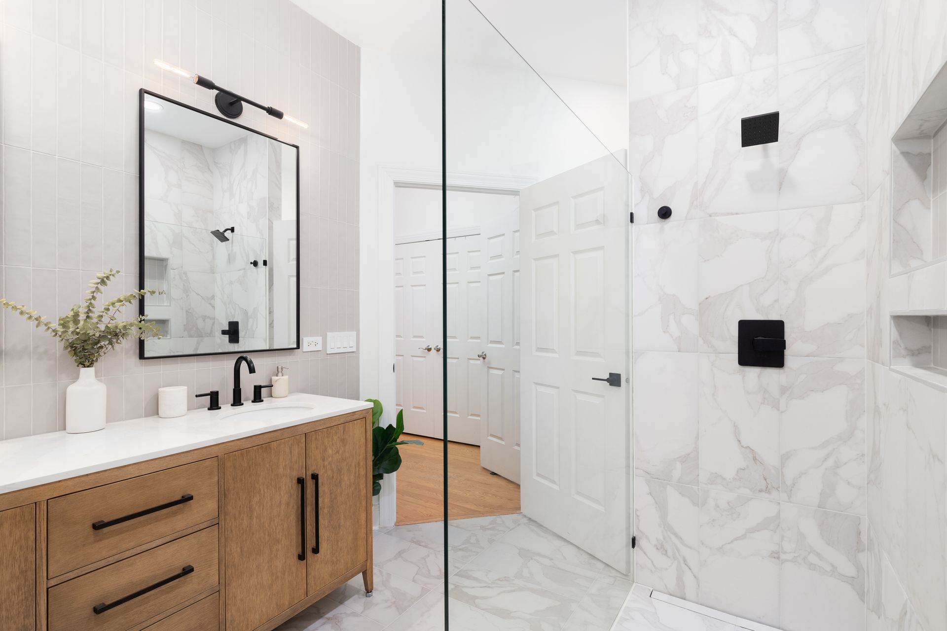 A modern bathroom with a wood-tone vanity, marble-tiled shower, black fixtures, and a rectangular mirror.