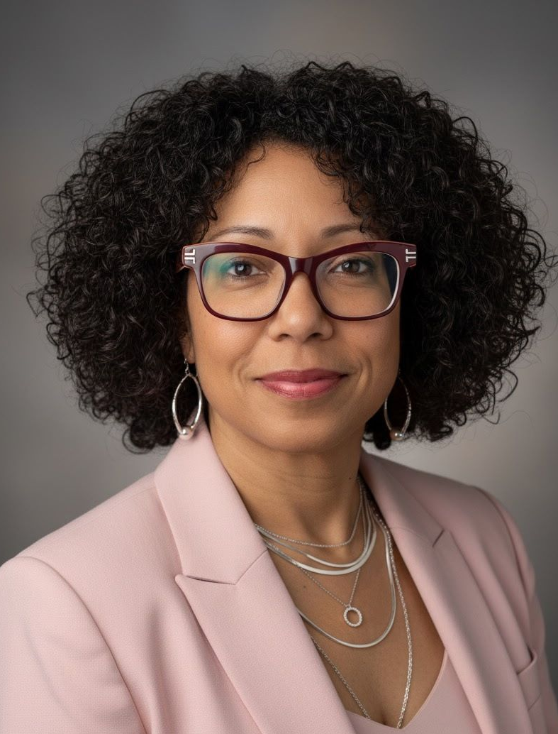 Woman with curly hair and glasses wearing a pink blazer, looking at the camera.