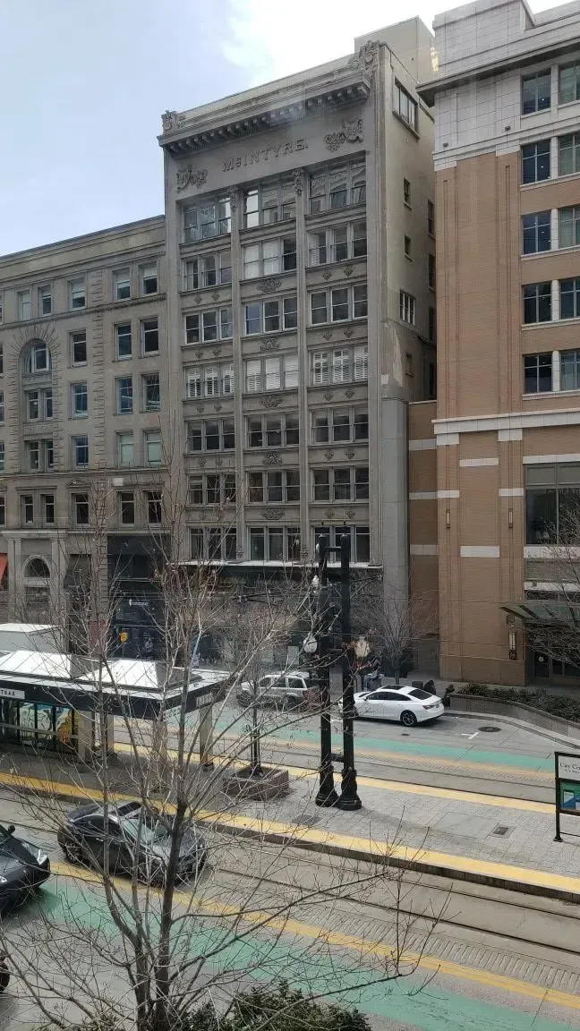 A street-level view of a city block featuring a tall, gray stone building next to a modern tan facade, with a light rail.