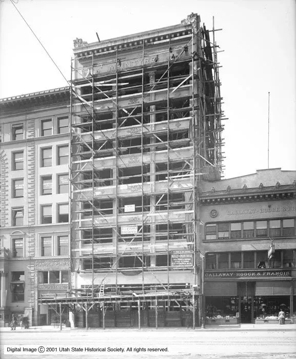A black-and-white photograph of a narrow building under construction, covered in wooden scaffolding, between two buildings.