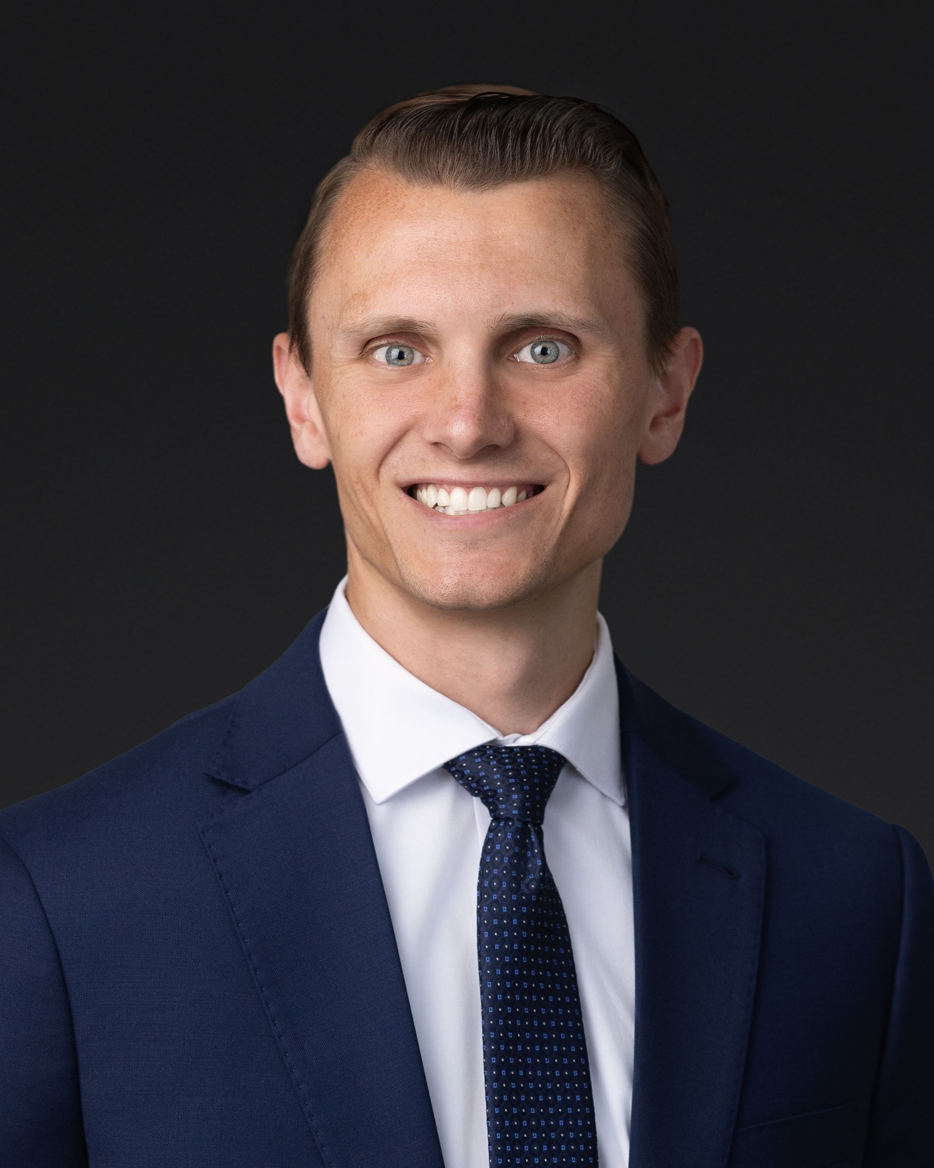 Studio headshot of a smiling man in a navy suit and tie against a dark background
