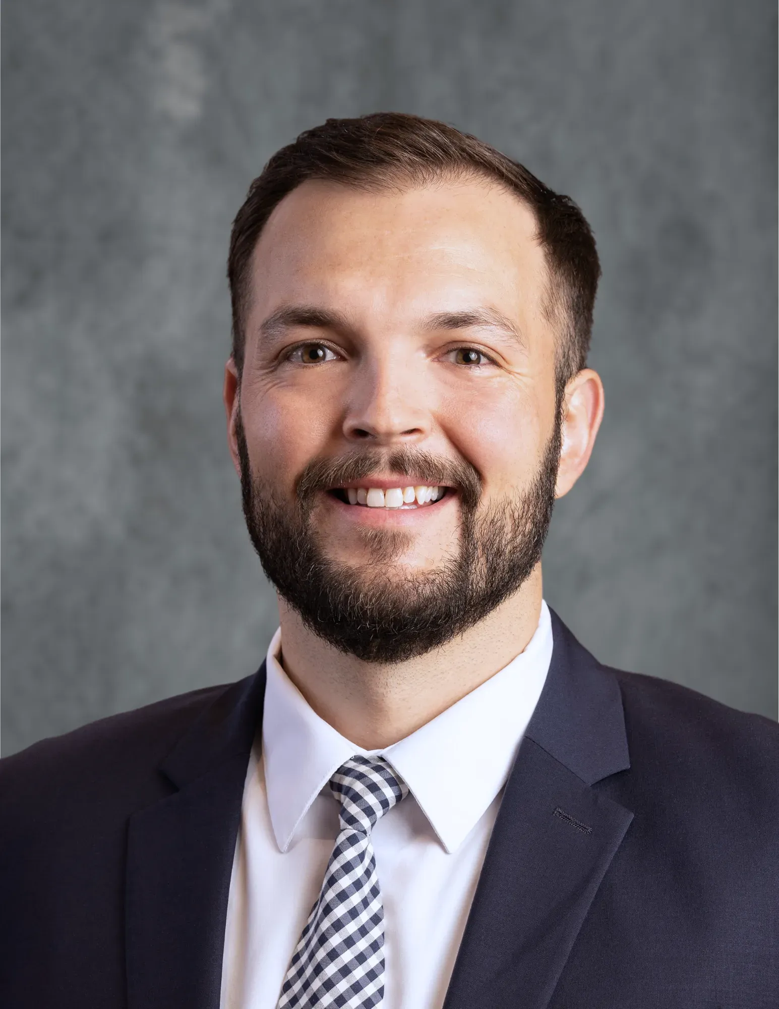 Man in a suit smiles, looking at the camera. He has a beard and a patterned tie against a gray backdrop.