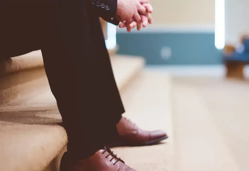 Person seated on carpeted steps, hands clasped, wearing dark pants and brown shoes.