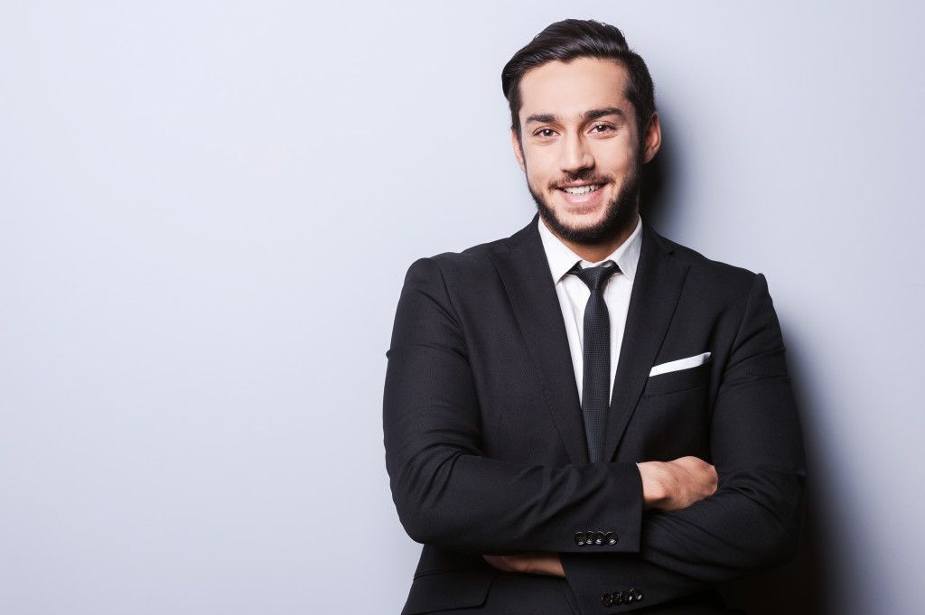 Man in a black suit standing with arms crossed against a light gray background, smiling.