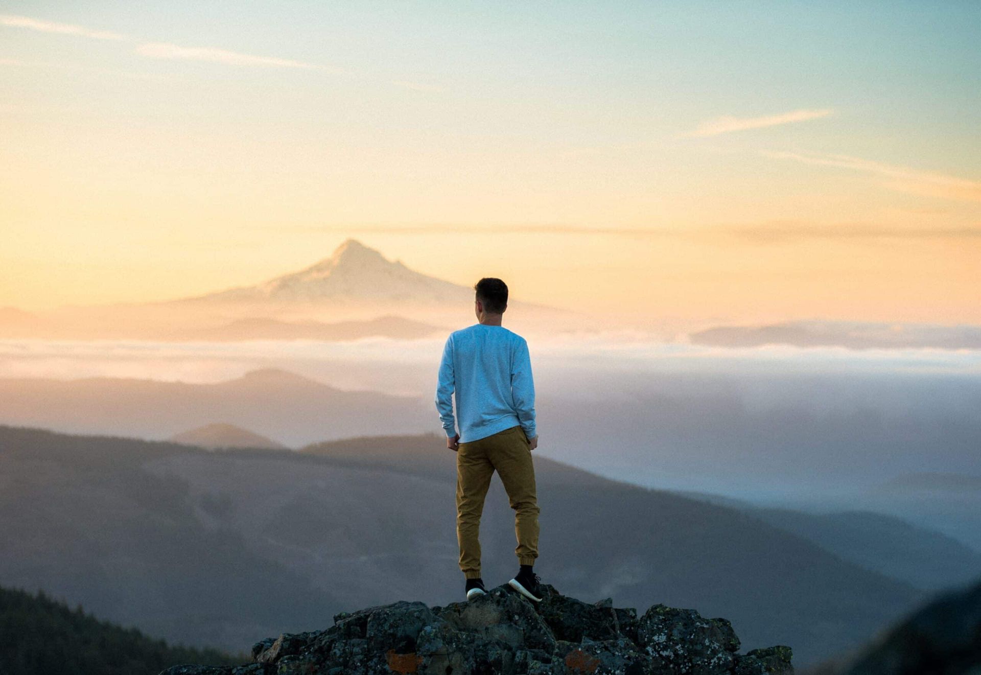 Person standing on a rocky peak, overlooking misty mountains at sunrise with a distant volcanic silhouette.