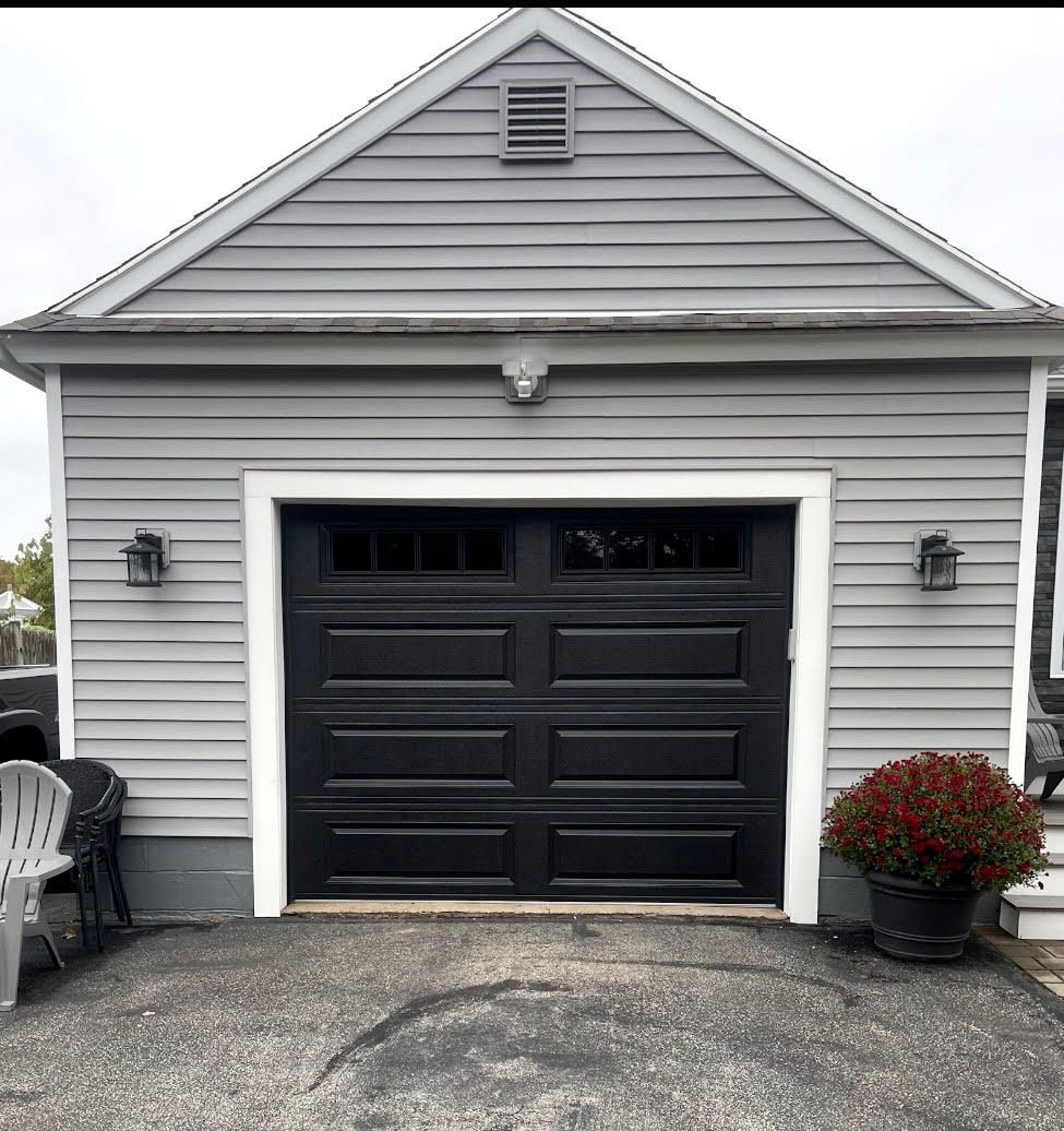 Black garage door on a gray-sided building with white trim; potted plant on right.