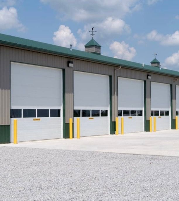 Exterior view of a building with four large white closed garage doors, yellow bollards, and a gravel surface.