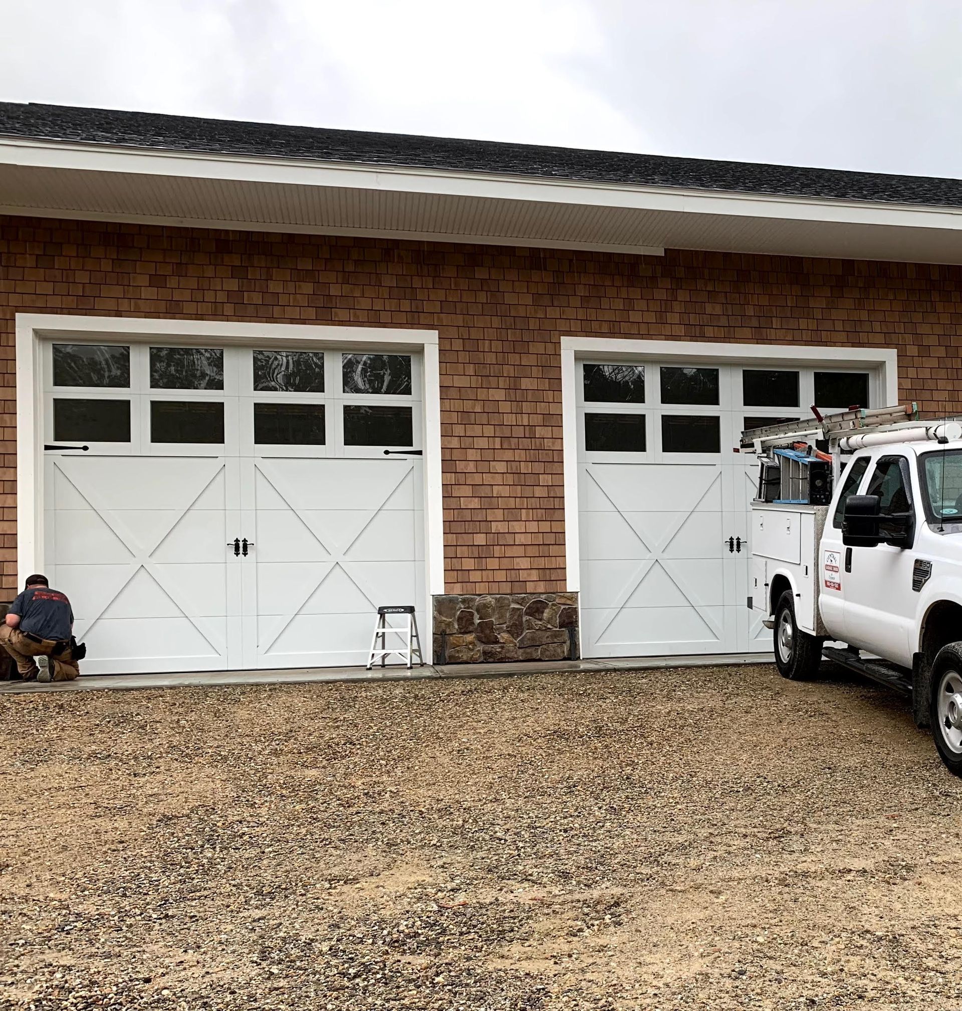 Two white garage doors on a brick building with a white truck parked next to the right door. A person is working on the left door.