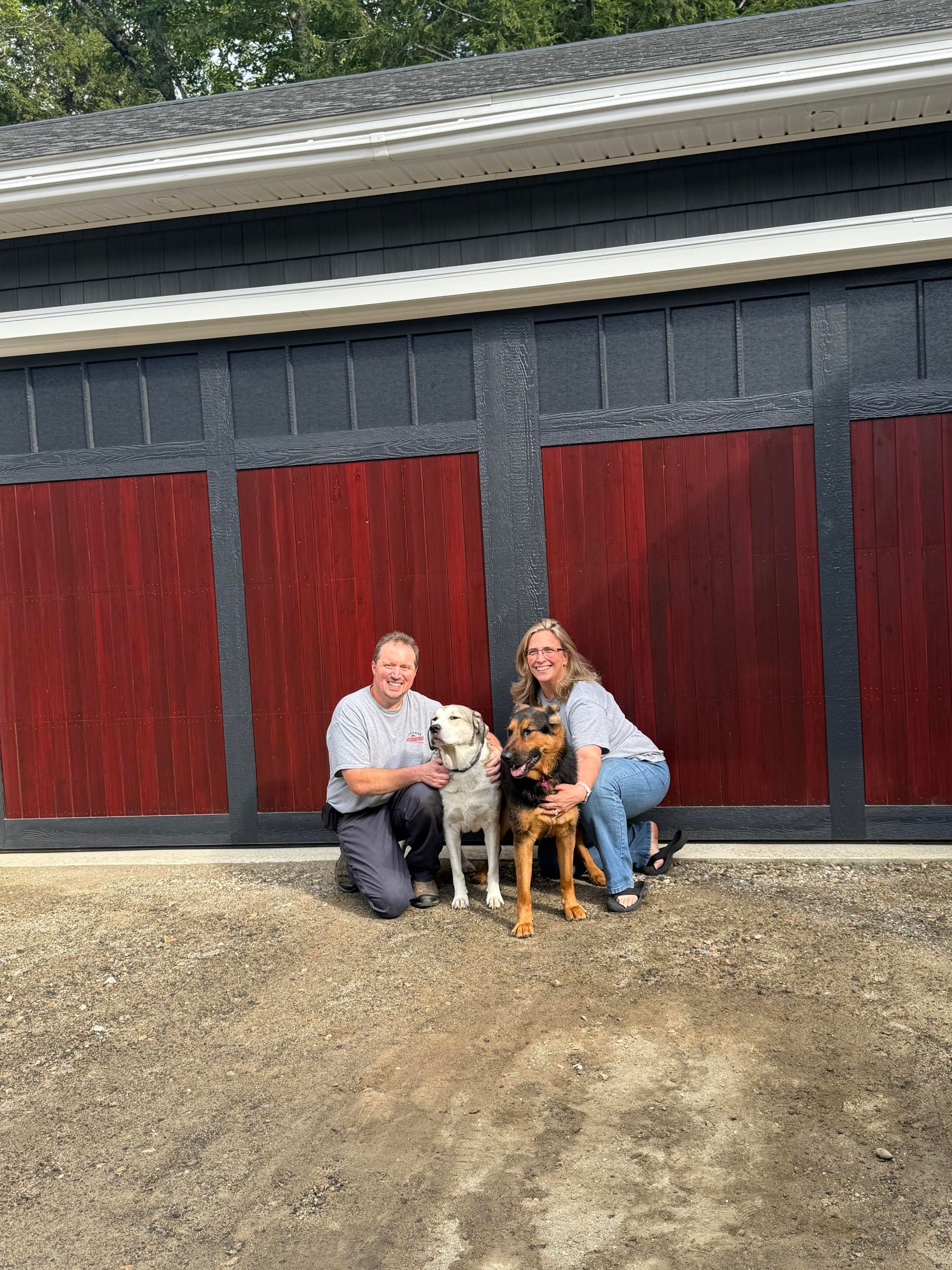 Two people kneeling with two dogs in front of a barn with red doors.