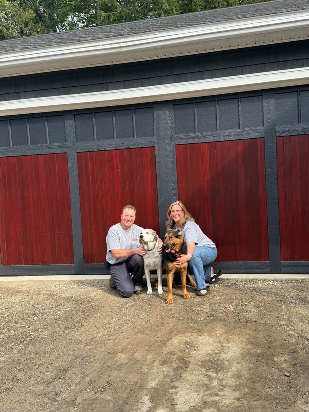 Two people kneel with two dogs in front of a barn with red doors.