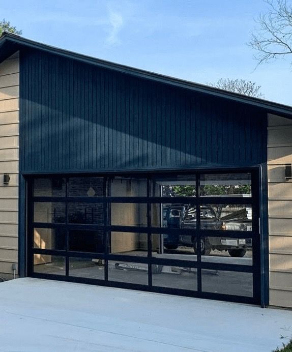 Modern garage with black-framed, clear glass door. Navy blue siding above. Tan siding on the sides.