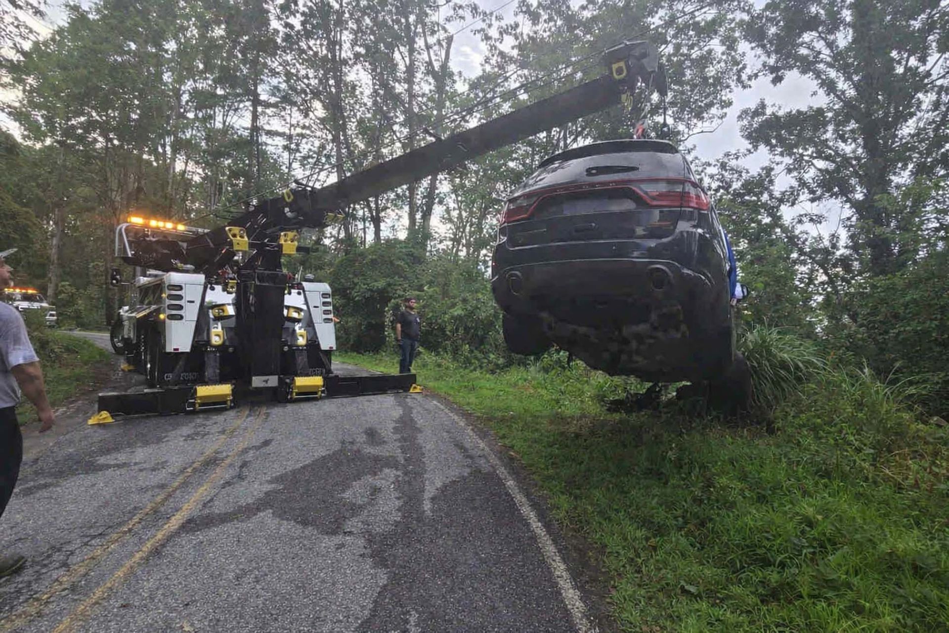 Tow truck lifting a black car off a wet roadside beside trees after a crash