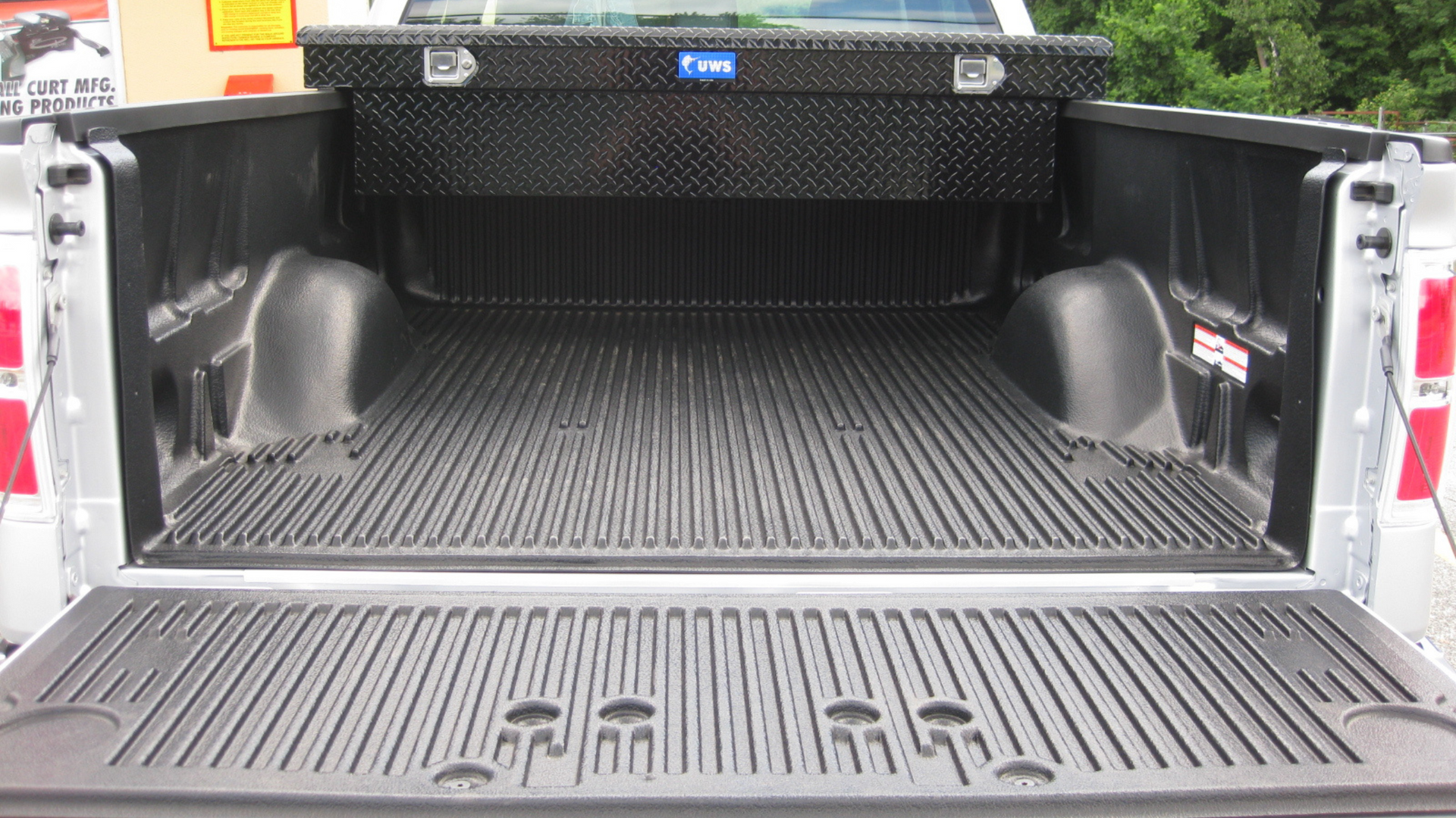 A truck bed with a black toolbox, lined with a black rubber mat. The tailgate is down.