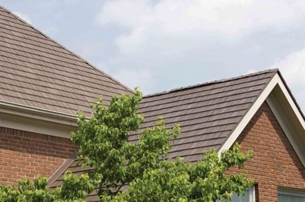Brown shingled roof of a brick house with a green tree and a cloudy sky.
