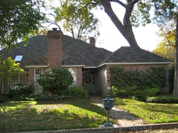 Brick house with dark roof, surrounded by green lawn and trees. Mailbox on sidewalk.