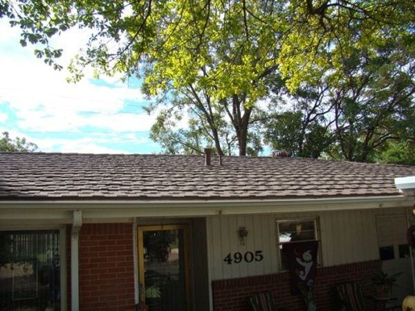 House exterior with brown shingle roof, brick facade, and trees against a partly cloudy sky.