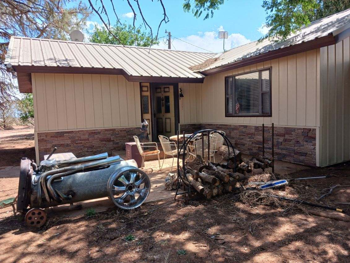 House exterior with light siding, a metal roof, and stone-like detail. An antique machine and firewood are outside.