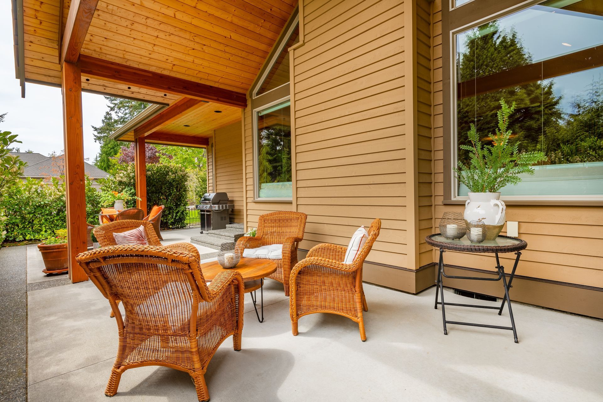 Covered patio with wicker seating, small table, and potted decor beside a beige house wall