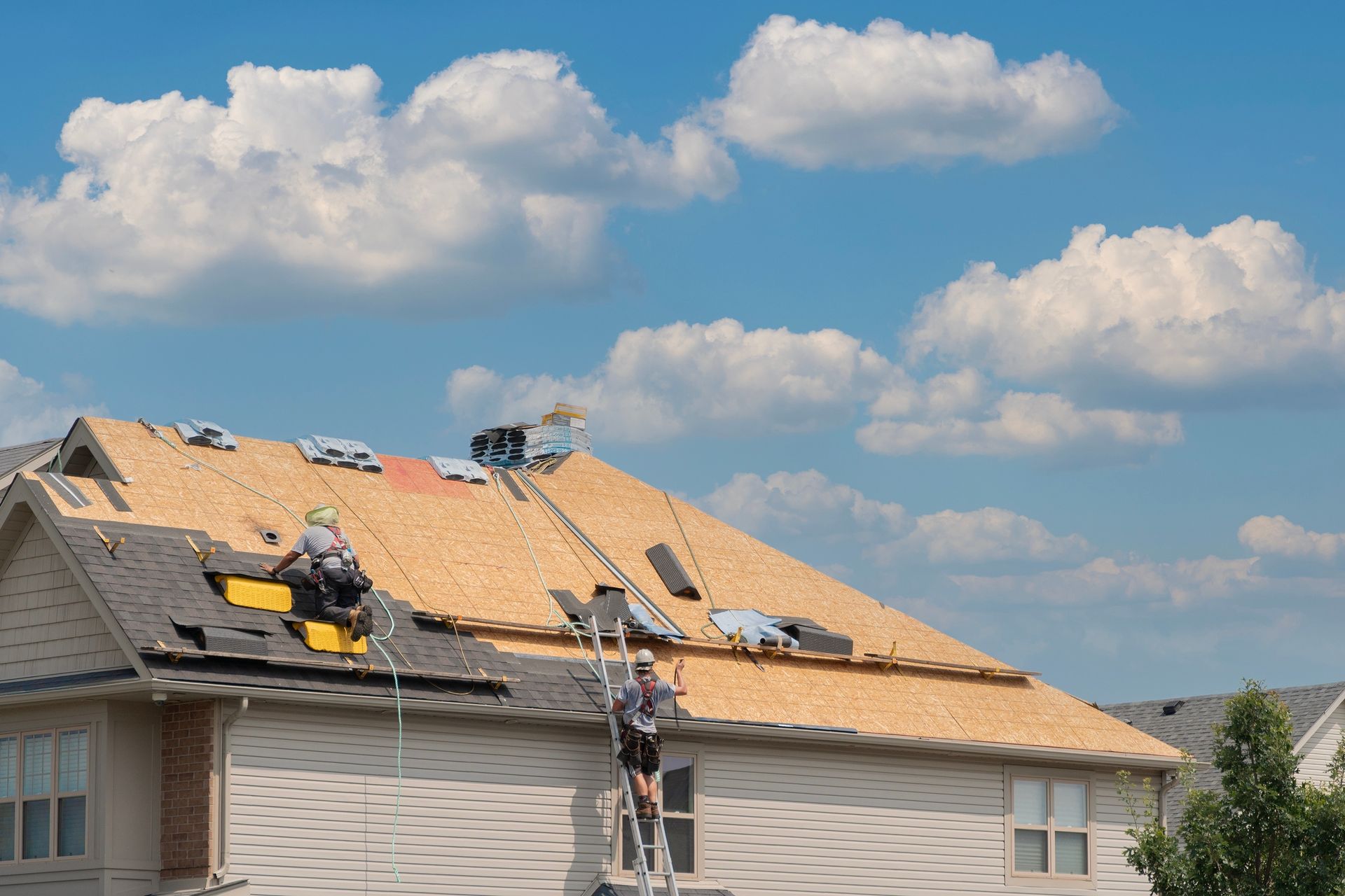 Workers installing shingles on a house roof under a blue sky with clouds