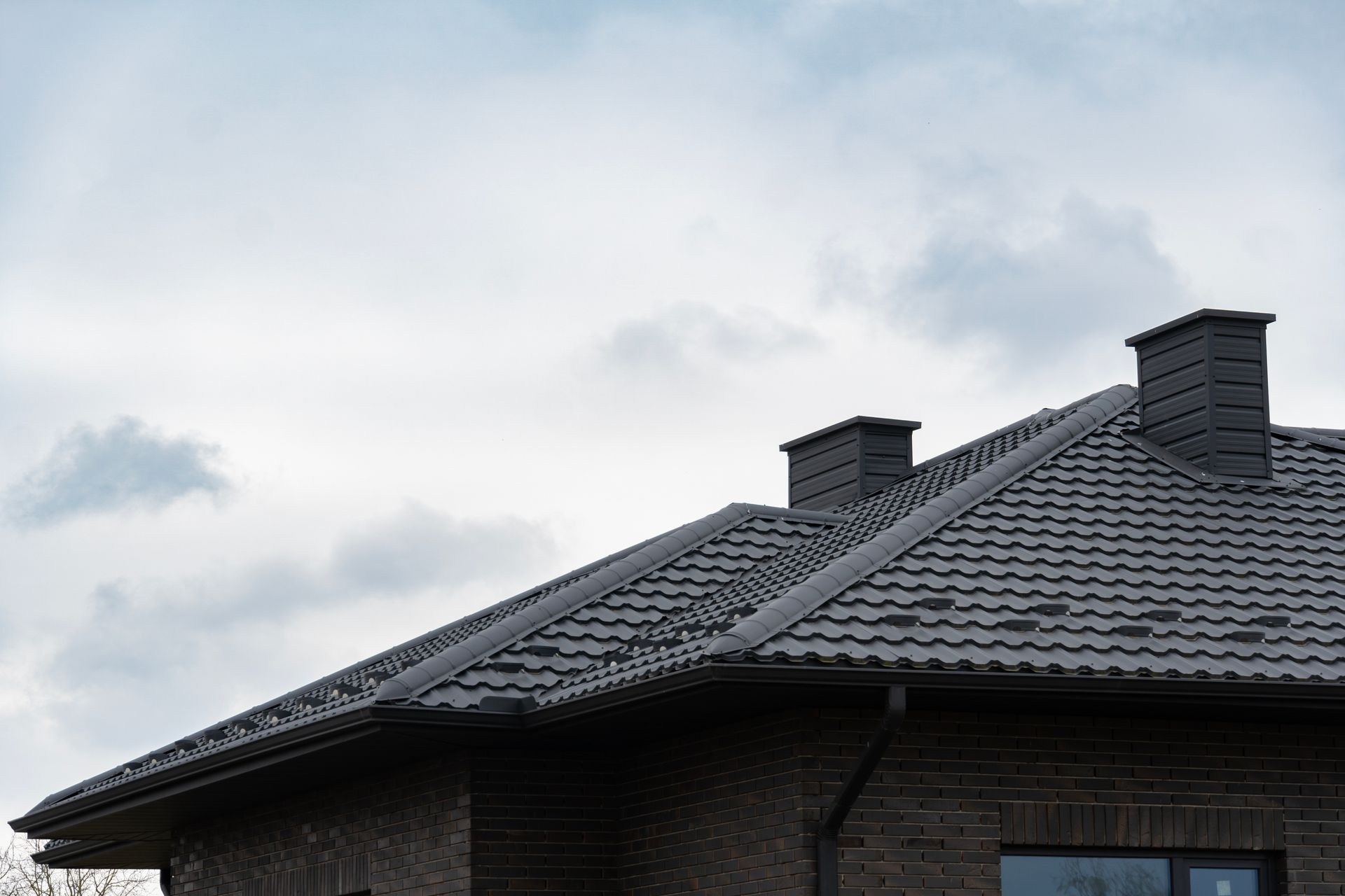 Dark gray tiled roof with two chimneys against a cloudy sky