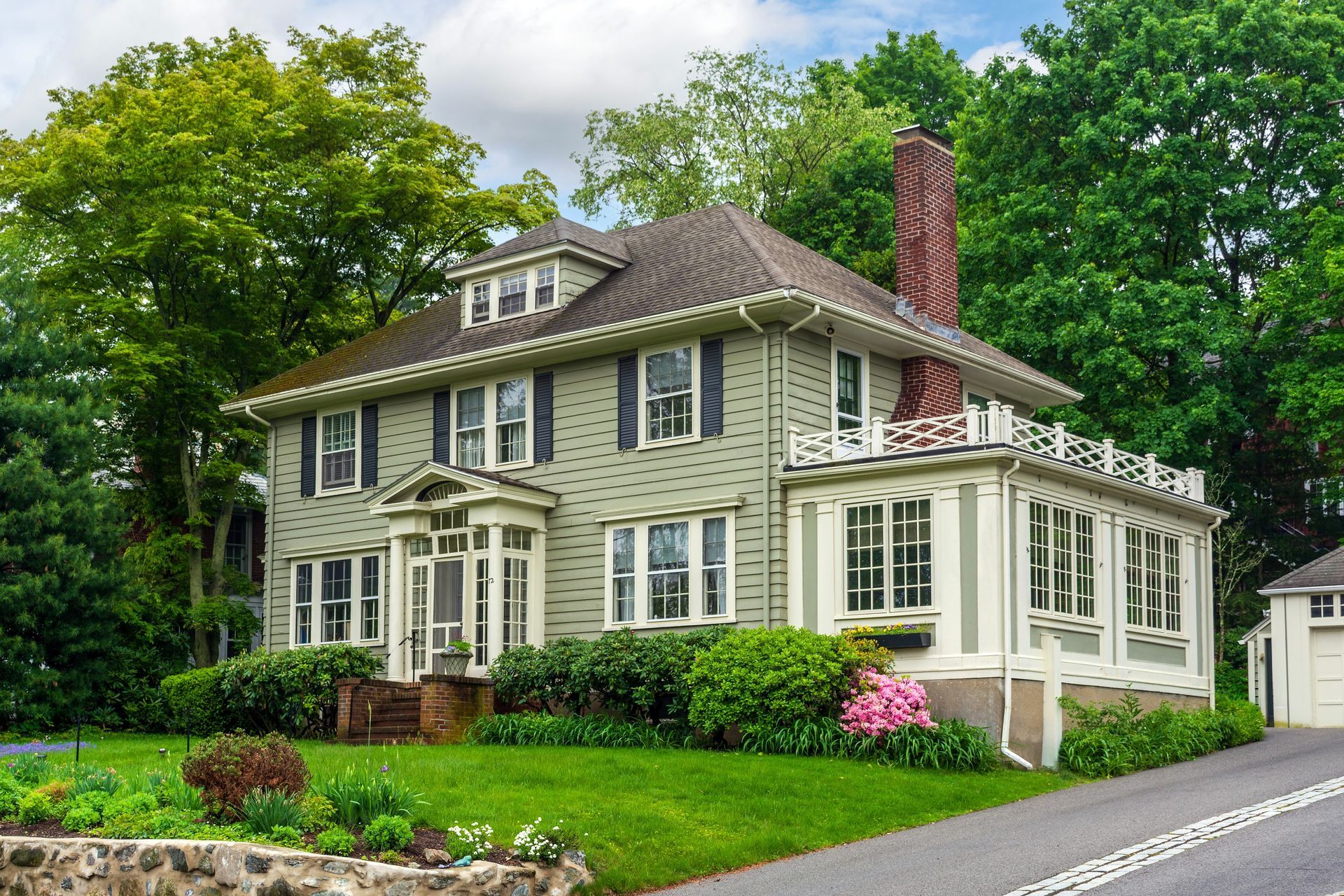 Large two-story green house with white trim, screened porch, red chimney, and lush trees by the road